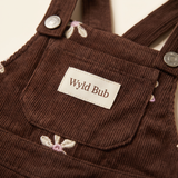Close-up of the cocoa corduroy pinafore bib showing the Wyld Bub woven label and embroidered flowers