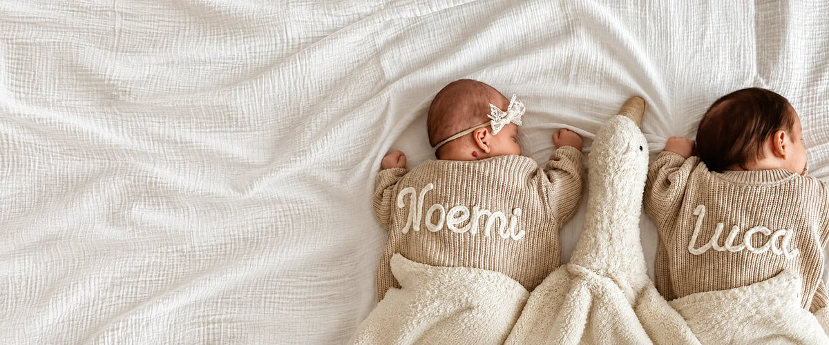 Two babies wearing personalized oatmeal sweaters with a cream cuddle duck, on ivory linen.